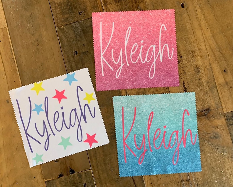 Op de afbeelding: Drie gepersonaliseerde stoffen vierkanten met de naam "Kyleigh" geschreven in verschillende stijlen. Het eerste vierkant heeft een witte achtergrond met kleurrijke sterren. Het tweede vierkant heeft een roze glitterachtergrond. Het derde vierkant heeft een blauwe en groene ombre glitterachtergrond.