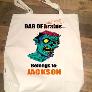 May include: White canvas tote bag featuring a zombie graphic and the text "BAG OF brains" with "treats" crossed out. The text "Belongs to: JACKSON" is printed in orange below the graphic.