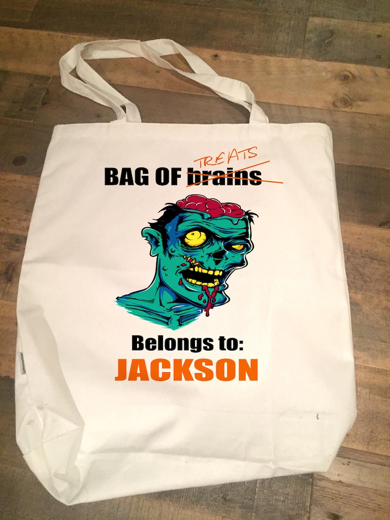 May include: White canvas tote bag featuring a zombie graphic and the text "BAG OF brains" with "treats" crossed out. The text "Belongs to: JACKSON" is printed in orange below the graphic.