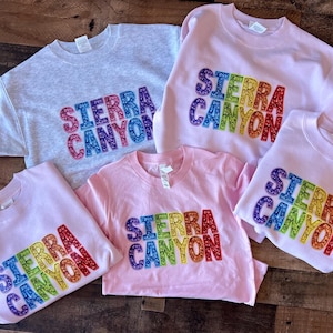 Könnte beinhalten: Vier Sweatshirts mit dem Text "SIERRA CANYON" in Regenbogen-Glitzerbuchstaben. Die Sweatshirts sind in verschiedenen Größen und Farben erhältlich, darunter Grau und Rosa.