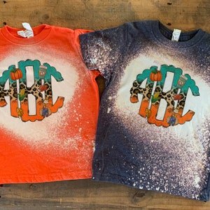 May include: Two short-sleeved t-shirts, one orange and one navy blue, with a bleached design. Each shirt features a circular graphic with the letters ABC, a pumpkin, and fall-themed illustrations. The shirts are on a wooden surface.