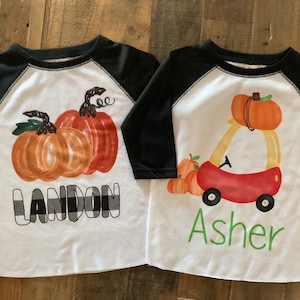 May include: Two white and black baseball shirts with a fall theme. One shirt has a pumpkin design with the name Landon in black and white plaid. The other shirt has a red car with pumpkins on top and the name Asher in green.