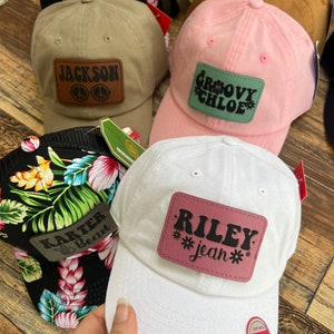 Könnte beinhalten: Vier verschiedene Baseballkappen mit personalisierten Patches. Die Kappen sind in verschiedenen Farben, darunter Schwarz, Weiß, Rosa und Braun. Die Patches zeigen verschiedene Namen und Designs, darunter "Jackson", "Groovy Chloe", "Karter Beau" und "Riley jean".