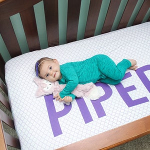 May include: A baby in a teal onesie lies on a crib sheet with the name "PIPER" in large purple letters. The sheet has a white and gray pattern. The baby is holding a pink stuffed animal. The crib is made of wood.