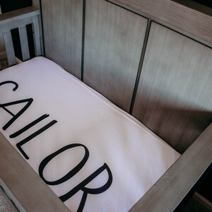 May include: A wooden crib with a white mattress featuring the word "SAILOR" in large, black letters. The crib has a light gray finish and is empty, ready for a baby.