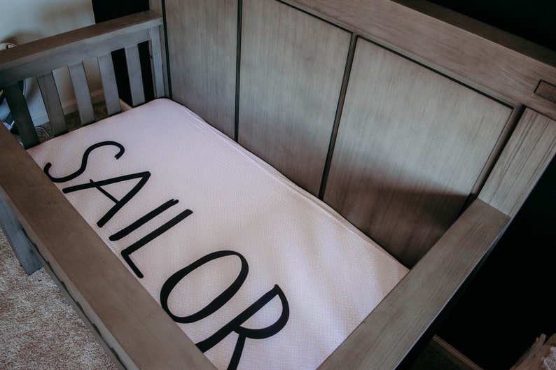 May include: A wooden crib with a white mattress featuring the word "SAILOR" in large, black letters. The crib has a light gray finish and is empty, ready for a baby.