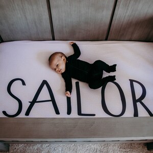 May include: A baby in a black onesie lies on a crib mattress. The mattress is white with the name "SAILOR" printed in large, black letters. The crib is made of gray wood.