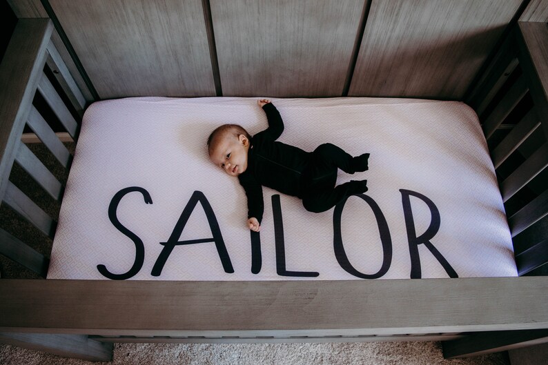 May include: A baby in a black onesie lies on a crib mattress. The mattress is white with the name "SAILOR" printed in large, black letters. The crib is made of gray wood.