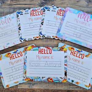 May include: Six colourful name tags with a "Hello" greeting and a line for writing a name. Each tag has a different design featuring cartoon cars, dinosaurs, and stars.