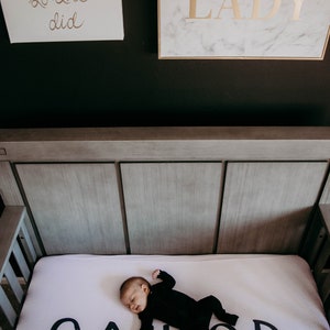 May include: A baby in a black onesie sleeps on a crib mattress with the word "SAILOR" in large black letters. Above the crib are two framed wall art pieces with gold lettering on white backgrounds. One reads "believed she could so she did" and the other says "BOSS LADY".