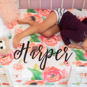 May include: A crib with a floral patterned sheet featuring large watercolor-style flowers in shades of pink, orange, and green. The name "Harper" is written in black script across the sheet. A child is sleeping on the sheet.