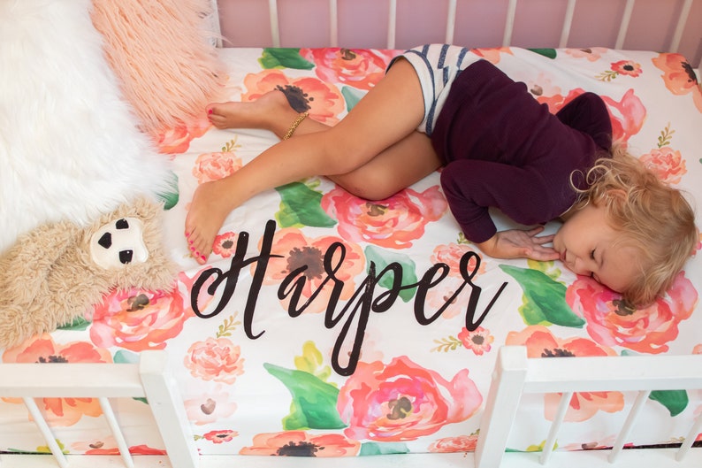 May include: A crib with a floral patterned sheet featuring large watercolor-style flowers in shades of pink, orange, and green. The name "Harper" is written in black script across the sheet. A child is sleeping on the sheet.
