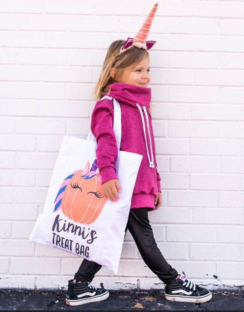 May include: A young child wearing a pink sweatshirt and black leggings is holding a white trick-or-treat bag with a unicorn pumpkin design. The bag has the text "kinni's treat bag" printed on it. The child is wearing a unicorn headband with a pink horn.
