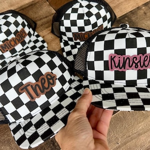 May include: Black and white checkered trucker hats with personalized names. The hats have a mesh back and a curved brim. The names "Michael", "Theo", and "Kinsley" are displayed on the front of the hats in a brown or pink font.