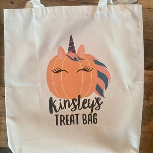 May include: White canvas tote bag featuring a unicorn pumpkin design and the text "Kinsley's Treat Bag".