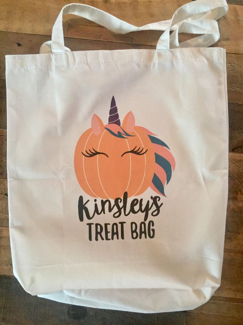 May include: White canvas tote bag featuring a unicorn pumpkin design and the text "Kinsley's Treat Bag".