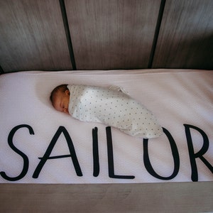 May include: A newborn baby swaddled in a white blanket with a small black dot pattern, sleeping peacefully in a crib. The crib mattress is light pink and features the name "SAILOR" in large, black letters.