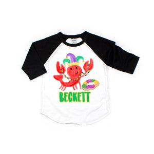 May include: White and black baseball tee shirt with a red cartoon lobster wearing a purple and green jester hat and beads. The lobster is holding a purple and green donut. The text "BECKETT" is printed in green on the shirt.