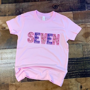 May include: Pink t-shirt with the word "SEVEN" in sparkly pink, purple, and red letters.
