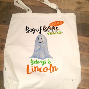 May include: White canvas tote bag featuring a ghost illustration and the text "Bag of Boo's Treats" with "Tricks" crossed out. The text "Belongs to: Lincoln" is written in orange below the ghost.