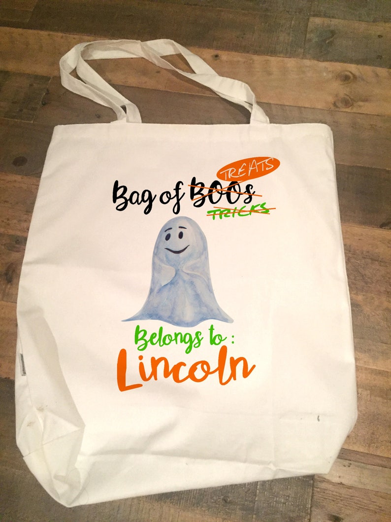 May include: White canvas tote bag featuring a ghost illustration and the text "Bag of Boo's Treats" with "Tricks" crossed out. The text "Belongs to: Lincoln" is written in orange below the ghost.