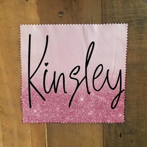 Op de afbeelding: Een vierkant van roze en wit ombre stof met de naam "Kinsley" geschreven in zwarte cursieve letters. De stof heeft een glinsterende textuur.