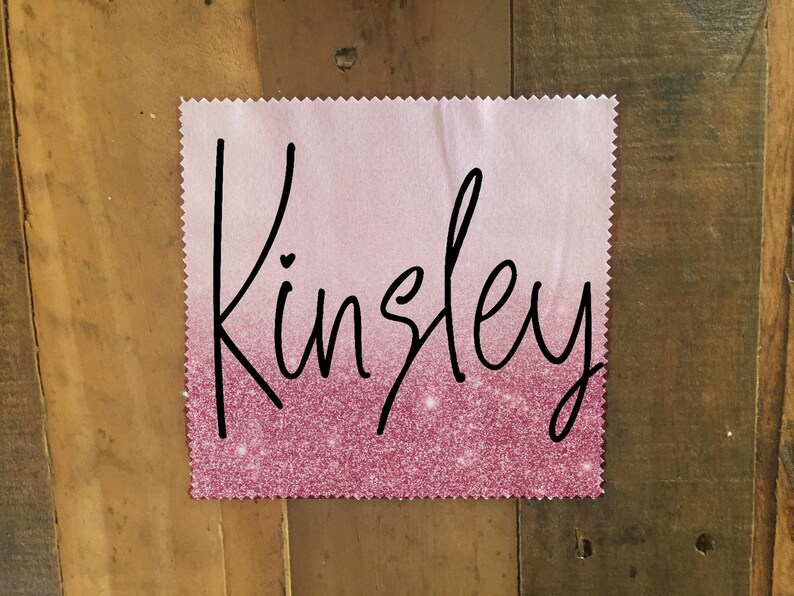 Op de afbeelding: Een vierkant van roze en wit ombre stof met de naam "Kinsley" geschreven in zwarte cursieve letters. De stof heeft een glinsterende textuur.