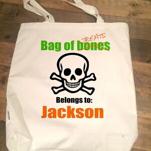 May include: White canvas tote bag with a black and white skull and crossbones design. The text "Bag of bones" is written in green and the text "Treats" is crossed out. The text "Belongs to: Jackson" is written in orange.