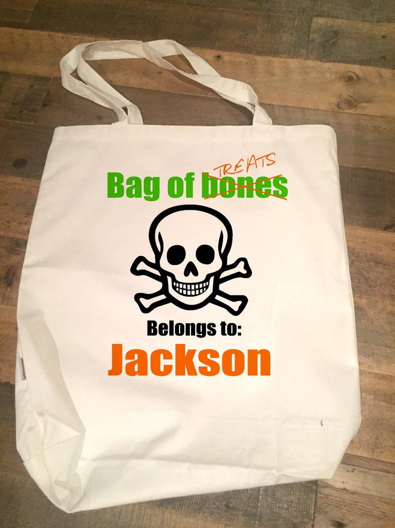 May include: White canvas tote bag with a black and white skull and crossbones design. The text "Bag of bones" is written in green and the text "Treats" is crossed out. The text "Belongs to: Jackson" is written in orange.