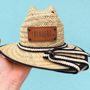 Puede incluir: Sombrero de paja con un parche de cuero marrón que dice "THEODORE". El sombrero tiene una banda a rayas blancas y negras y una correa de barbilla negra. El sombrero está hecho de paja tejida y tiene un ala pequeña.