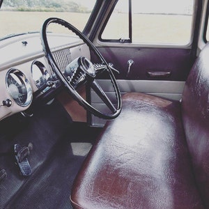 May include: Interior shot of a classic truck. The image focuses on the driver's side, featuring a black steering wheel, brown leather seat, and dashboard with gauges. The interior has a retro style, highlighting the vehicle's design details.