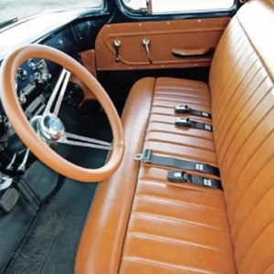 May include: Interior of a classic car with a tan leather bench seat and black seatbelts. The steering wheel is tan, and the dashboard is black with chrome details. The car's interior has a vintage design, with a classic aesthetic.
