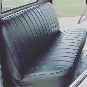May include: Black vinyl bench seat with vertical ribbed detailing. The seat is within a vehicle, with a window visible in the background. The seat has a curved design and appears to be in good condition.