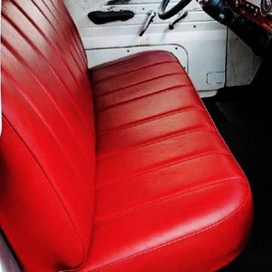 以下が含まれることがあります： 鮮やかな赤色の車のベンチシートのクローズアップ。シートは、水平にステッチされたラインを持つ、滑らかな革のような素材で作られています。車の内装は白で、黒いハンドルとドアハンドルが見えます。