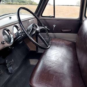 May include: Interior view of a classic truck cab. The image shows a black steering wheel, brown leather bench seat, and a dashboard with gauges. The interior has a vintage aesthetic, with a focus on the vehicle's design and materials.