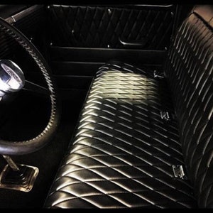 Puede incluir: Interior de coche clásico con tapicería de cuero negro acolchada con diseño de rombos. El volante cromado se ve a la izquierda. El diseño es vintage, con una estética oscura y evocadora. El interior del coche está bien conservado.