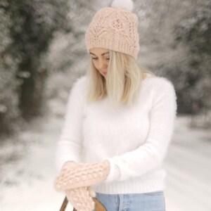 Puede incluir: Una mujer con un gorro de punto rosa con un pompón blanco, un suéter blanco peludo y guantes de punto rosa. Está de pie en un bosque nevado.