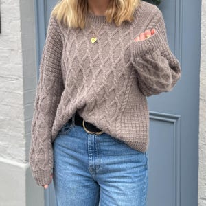 May include: A brown cable knit sweater with a gold heart necklace. The sweater has a relaxed fit and is worn with blue jeans and a black belt.