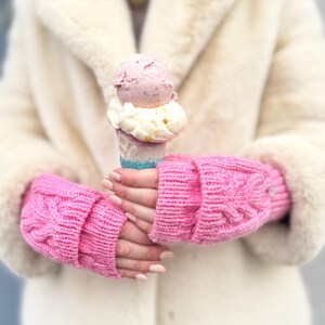 May include: A pair of pink knitted fingerless gloves with a cable knit pattern. The gloves are being worn by a person holding an ice cream cone with two scoops of ice cream.