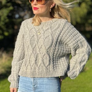 Könnte beinhalten: Ein grauer Strickpullover mit Zopfmuster, lockerer Passform und langen Ärmeln. Der Pullover hat einen runden Ausschnitt und eine verkürzte Länge.