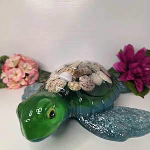 May include: A decorative turtle figurine with a green head and shell adorned with seashells. The body transitions from green to blue with glitter accents. Pink and purple flowers are in the background.