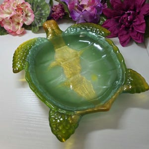 May include: A sea turtle shaped dish crafted from translucent green glass. The center of the shell features a prominent, textured gold-colored inlay that extends toward the head. The flippers and edges of the shell display a darker green hue.