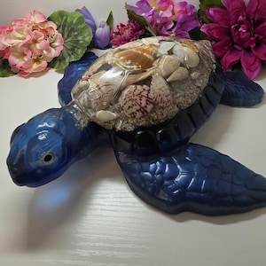 May include: A decorative sea turtle figurine featuring a deep blue translucent resin body and flippers. The shell is a clear, hollow dome filled with a layer of sand and an assortment of various small, natural-colored seashells.