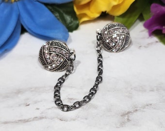 May include: A pair of silver tone, rhinestone encrusted, decorative clasps connected by a black chain.