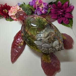 May include: A decorative sea turtle figurine crafted from translucent resin with a reddish-pink hue. The shell is hollow and contains a miniature diorama featuring sand, various small seashells, and green foliage visible through the clear top.