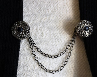 May include: Two silver and black floral brooches connected by a silver chain. The brooches have a black stone in the centre.