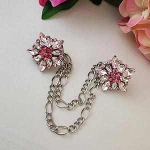 Puede incluir: Broche plateado con dos adornos en forma de flor unidos por una cadena. Cada flor presenta cristales rosas y transparentes. El broche está sobre una superficie blanca, con flores rosas en el fondo.