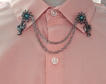 May include: A silver chain collar pin with a turquoise flower and cactus design. The pin is attached to a pink shirt.