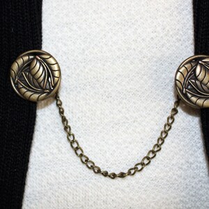 Puede incluir: Un par de botones de metal dorado con un dise&ntilde;o de hojas conectados por una cadena. Los botones est&aacute;n unidos a una chaqueta negra.