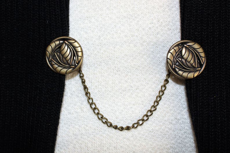 Puede incluir: Un par de botones de metal dorado con un dise&ntilde;o de hojas conectados por una cadena. Los botones est&aacute;n unidos a una chaqueta negra.